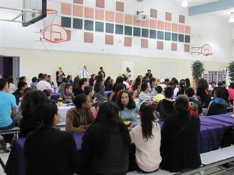 assembly of parents and students that have received honor roll awards sit at cafeteria tables eating.