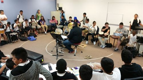 Students with various drums gathered in a circle around a teacher at a full drum set.